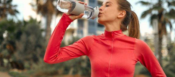 Young slender woman in sportswear with a bottle of water standing on the street against the backdrop of palm trees and buildings. Sports and Recreation.